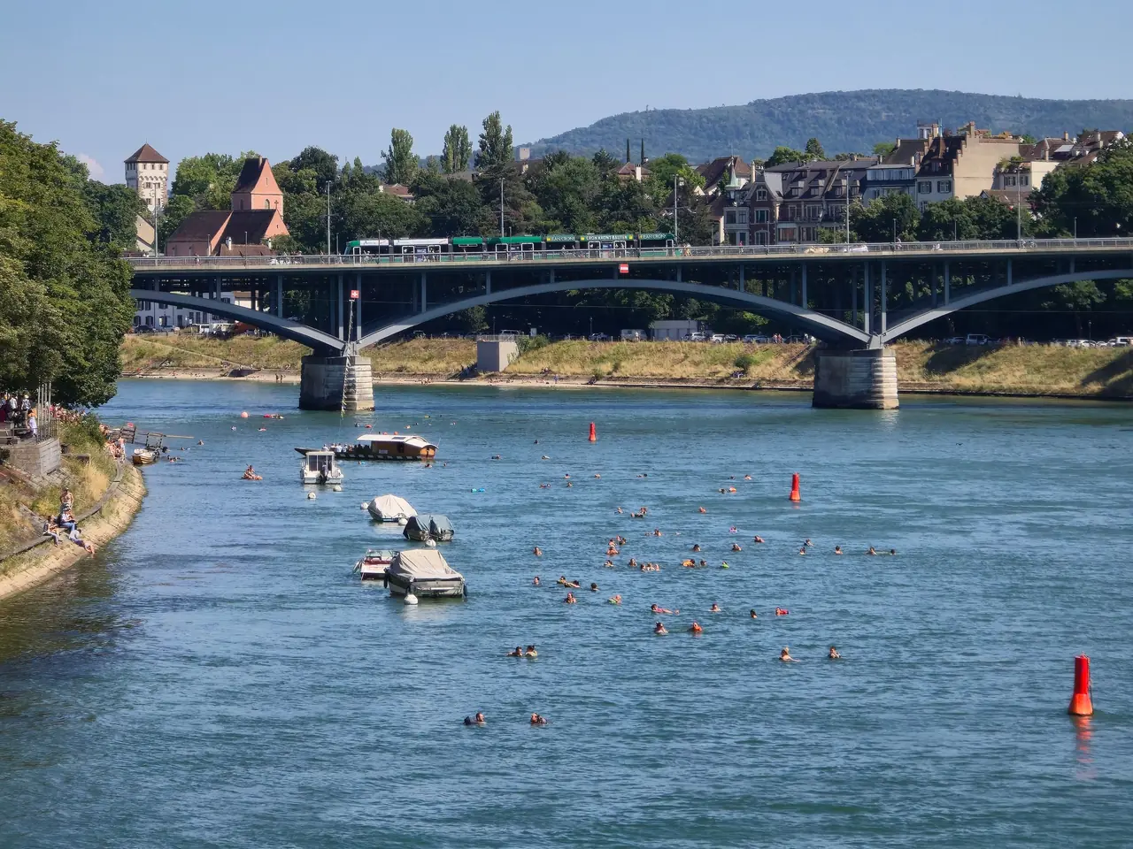 Des nageurs et des bateaux sur le Rhin à Bâle, devant un pont, un jour d’été.