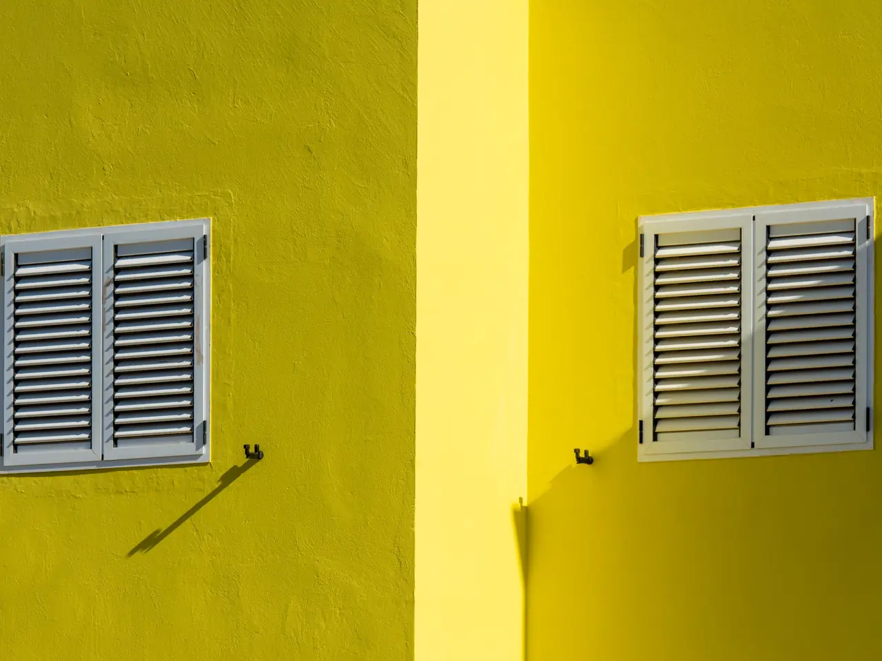 Deux fenêtres aux volets à persiennes fermés, sur une façade jaune en plein soleil.