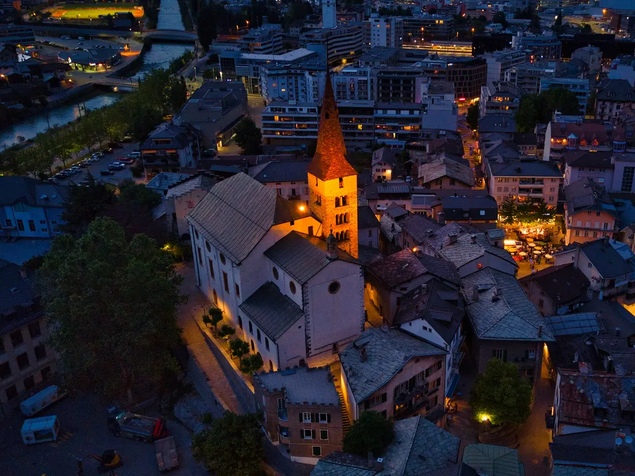 Viège, en Valais, vue du ciel à la nuit tombée : l’église éclairée au milieu des toits.